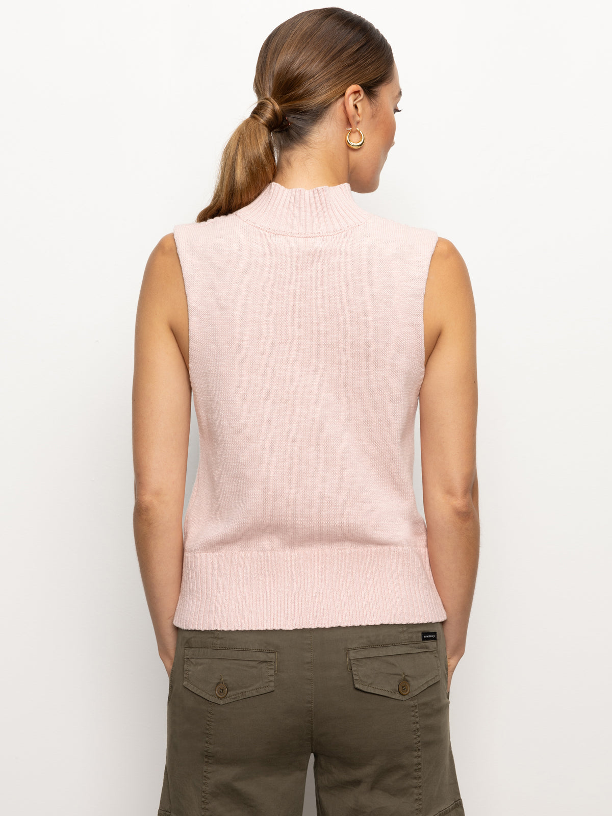 A woman with brown hair in a ponytail is wearing the rib detail mock tank in rosewood by Sanctuary Clothing, paired with olive green pants featuring button pockets, and is facing away from the camera against a plain white background.