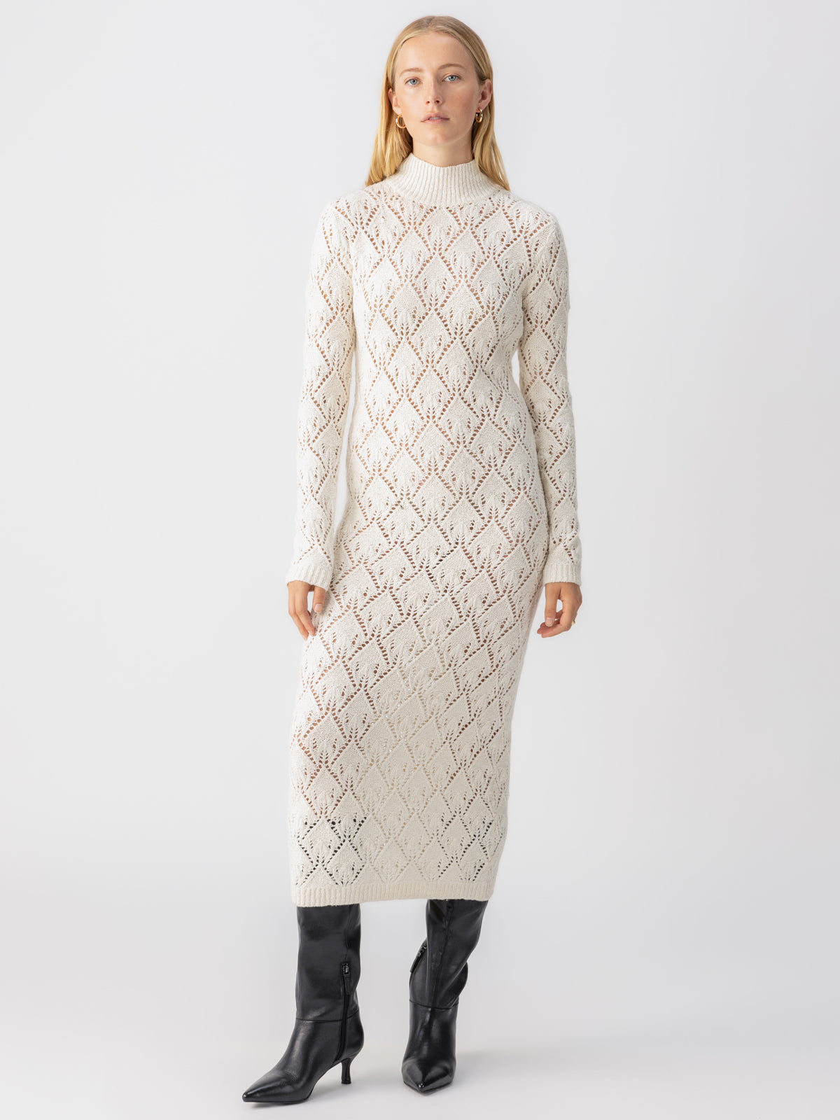 A woman stands against a plain background wearing the Pointelle Sweater Maxi Dress Eco Natural from Sanctuary Clothing, a long, fitted white knit dress with a high neckline and long sleeves. The dress features a delicate diamond pattern. She pairs the outfit with black knee-high boots. Her long, blonde hair is worn down.