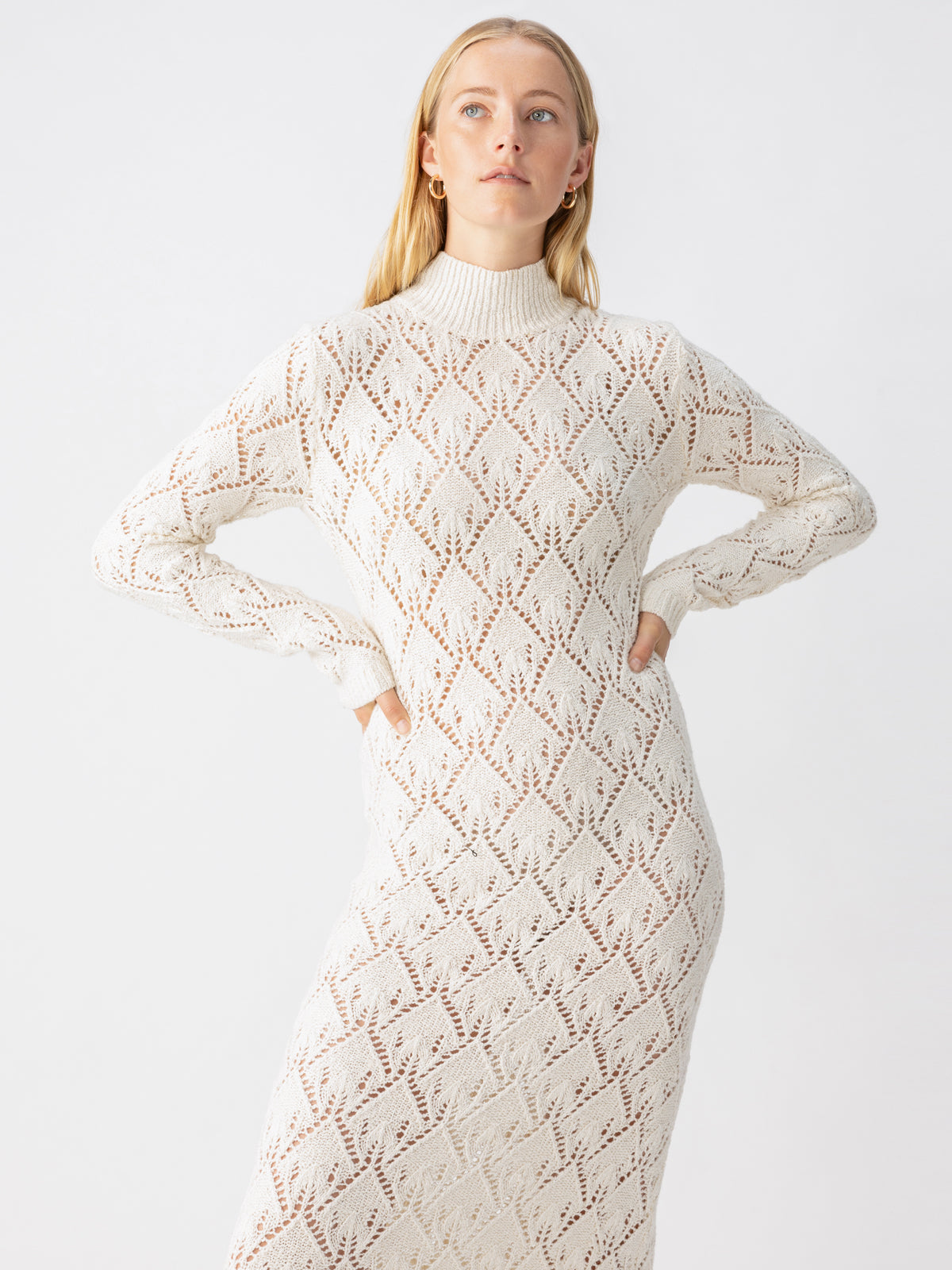 A person with long, blonde hair is wearing the Sanctuary Clothing Pointelle Sweater Maxi Dress Eco Natural, a white long-sleeved knit dress featuring a diamond pattern. They stand against a plain white background, with their hands on their hips, looking slightly to the side.