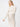 A person stands against a plain white background wearing the "Pointelle Sweater Maxi Dress Eco Natural" by Sanctuary Clothing. Their hair is down, and they have a serene expression, with one arm resting by their side and the other extended slightly outward.