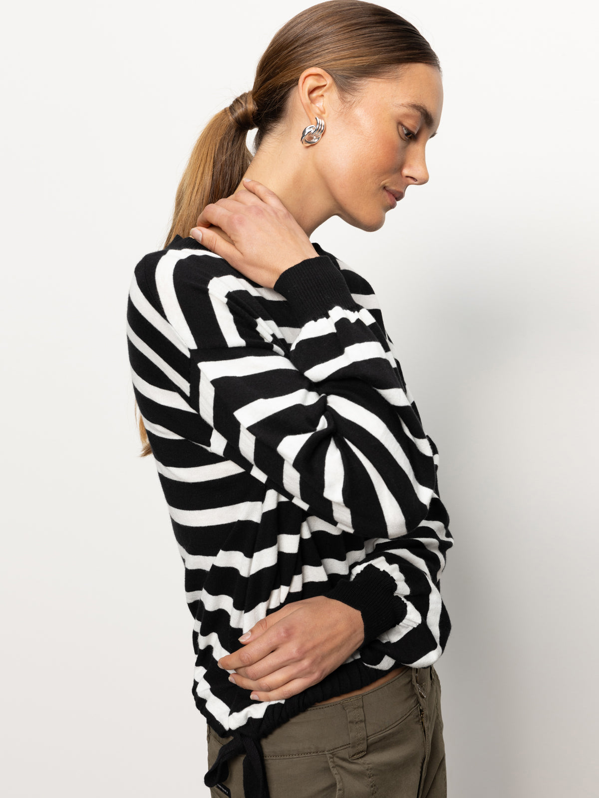 A person with long brown hair tied in a ponytail is wearing the Sanctuary Clothing side tie sweater in black chalk stripe. They are facing sideways, touching their neck with one hand against a plain white background.