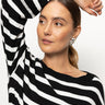 A person with their hair tied back wears the "side tie sweater blk chalk stripe" by Sanctuary Clothing, looking to the side with a relaxed expression. One arm is raised, resting on their head. They have hoop earrings and are against a plain background.