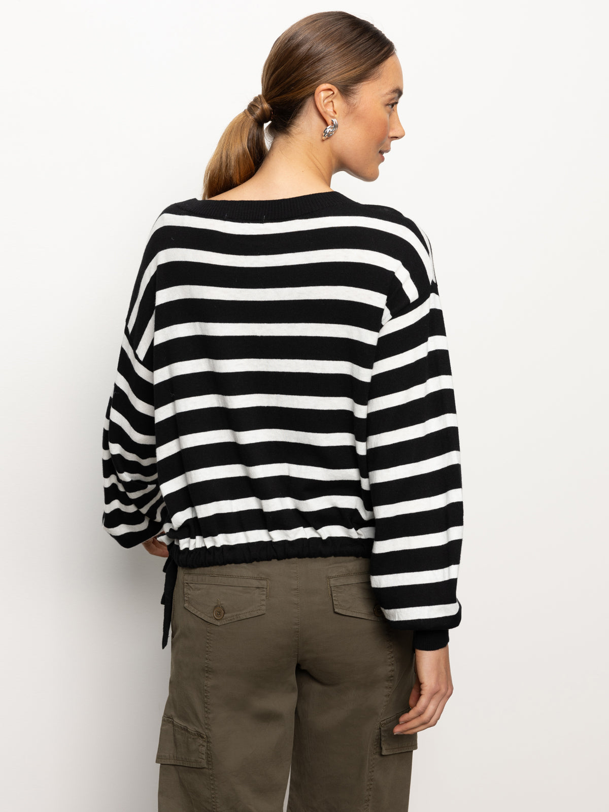 A person with long hair styled in a ponytail is wearing the "Side Tie Sweater" in black chalk stripe by Sanctuary Clothing, paired with olive green cargo pants. They are standing against a plain white background and facing away from the camera.