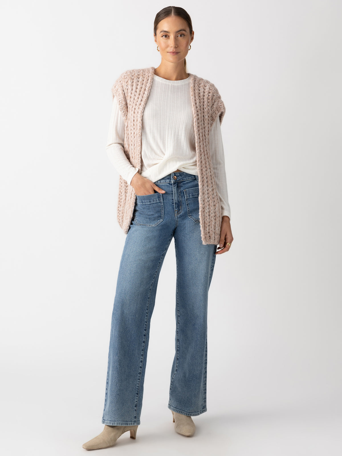 A person is standing against a plain background, wearing a chunky sweater vest in rosewood marl by Sanctuary Clothing over a white long-sleeve shirt, blue wide-leg jeans, and beige ankle boots. They have their hair pulled back and are looking at the camera.