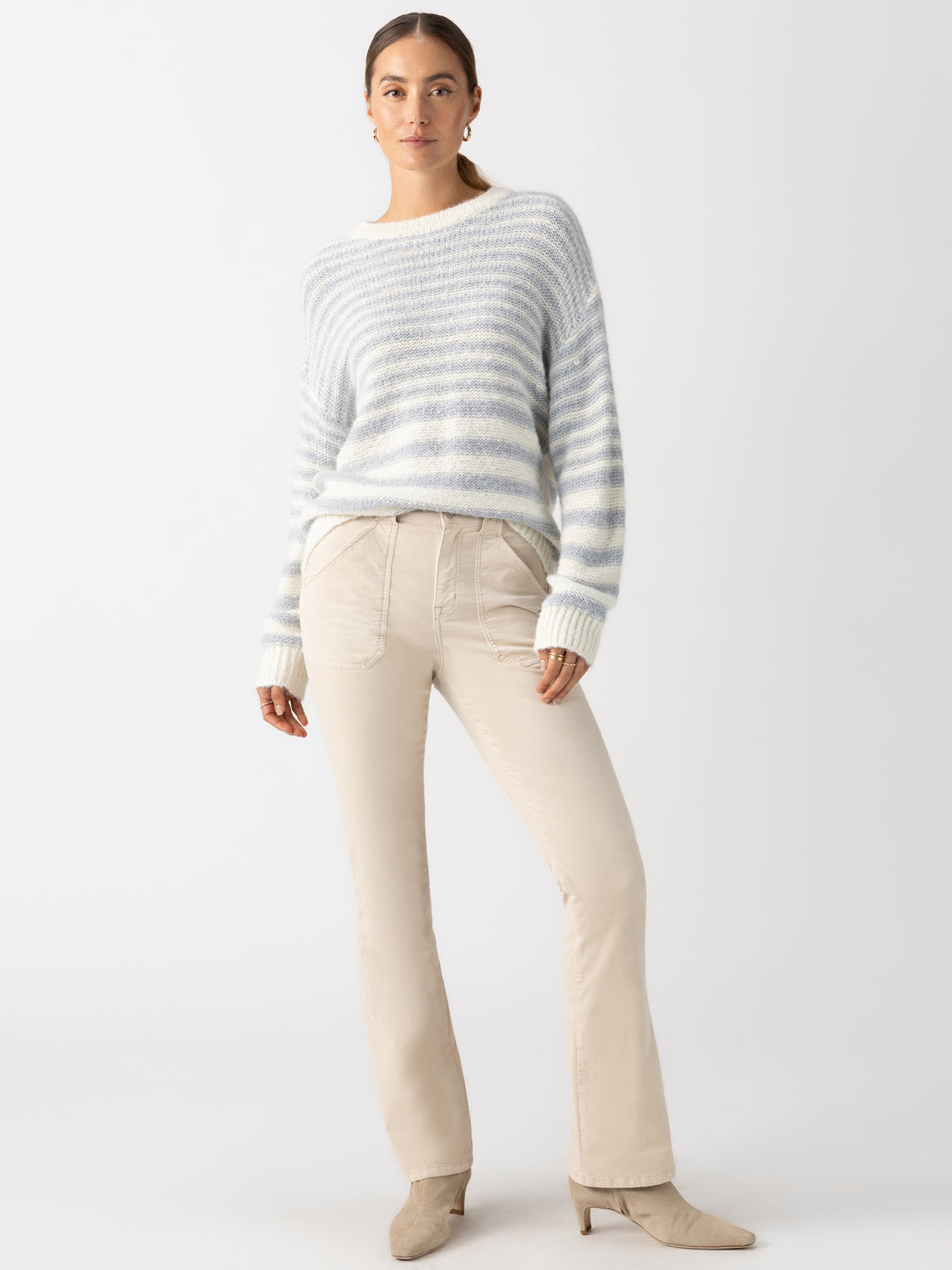 A person wearing a Sanctuary Clothing Fuzzy Tunic Sweater in Pearl Blue Chalk Stripe, along with beige pants and ankle boots, stands against a plain white background. They pose confidently with hands in pockets, looking at the camera.