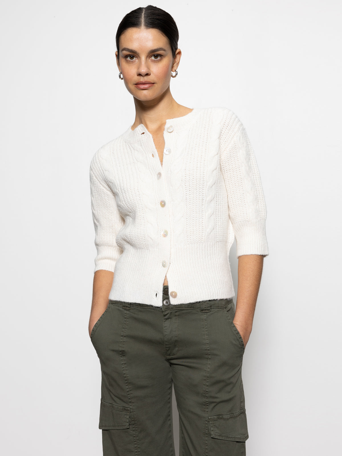 A person wearing Sanctuary Clothing's "fuzzy cable cardi light oat," with three-quarter sleeves, paired with olive green cargo pants, standing against a plain white background. They have short dark hair and are looking forward with hands in their pockets.