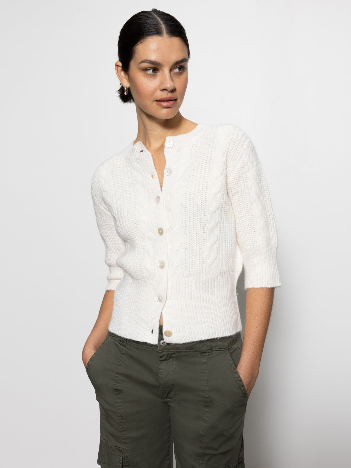 A person with dark hair tied back wears Sanctuary Clothing's "fuzzy cable cardi light oat," a textured white cardigan with a button-up front, paired with dark green pants. They stand against a plain white background, gazing to the side with hands in their pockets.