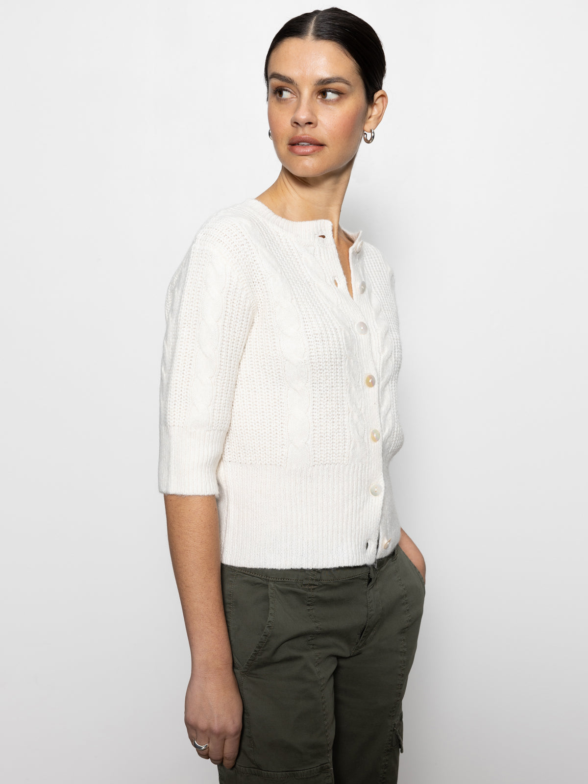 A person wearing an oat-colored, short-sleeved, button-up fuzzy cable cardigan from Sanctuary Clothing is standing against a plain white background. They are also dressed in olive green pants and small hoop earrings, gazing to their right.