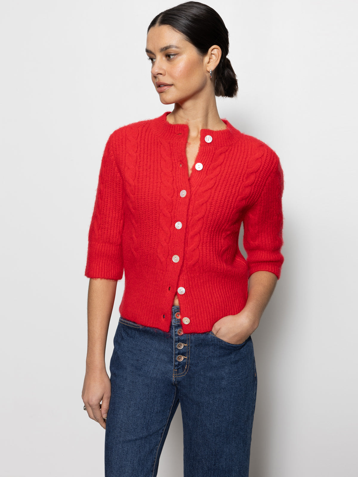 A person stands against a plain background wearing Sanctuary Clothing's "fuzzy cable cardi bittersweet," a vibrant red knitted cardigan featuring white buttons and elbow-length sleeves, paired with blue jeans. They have their hand in their jeans pocket and are looking to the side.