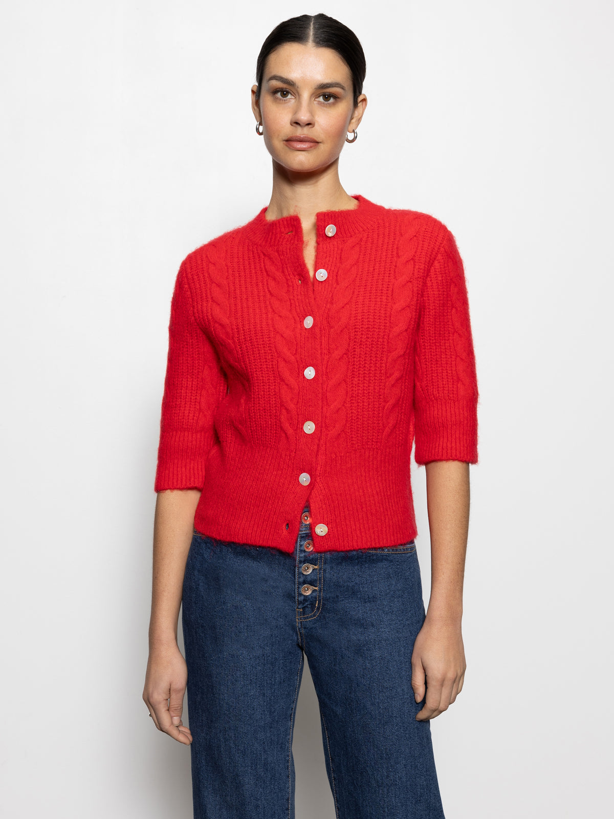 A person wearing the fuzzy cable cardi bittersweet by Sanctuary Clothing, a vibrant red, buttoned cardigan with three-quarter sleeves, and blue jeans stands against a plain white background.