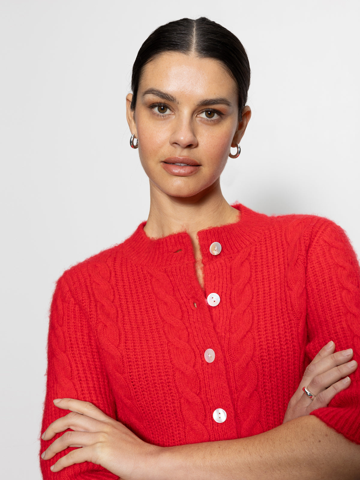 A person with dark hair is wearing the vibrant red "Fuzzy Cable Cardi Bittersweet" from Sanctuary Clothing, paired with silver hoop earrings. The background is plain and light-colored.