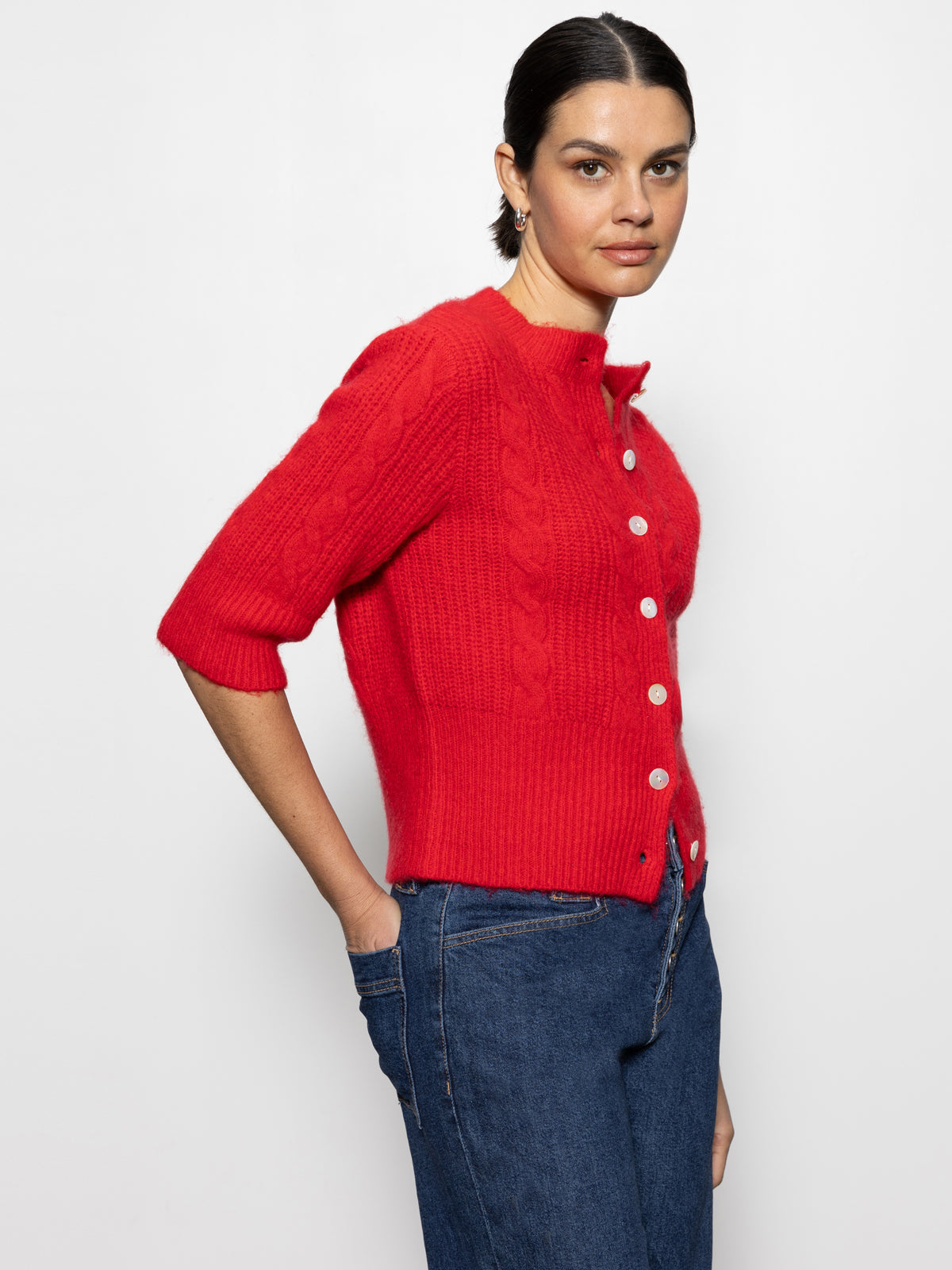 A person wearing the Sanctuary Clothing "fuzzy cable cardi bittersweet," a bright red cardigan with button-up design and elbow-length sleeves, paired with blue jeans stands against a plain white background, looking at the camera with a neutral expression.