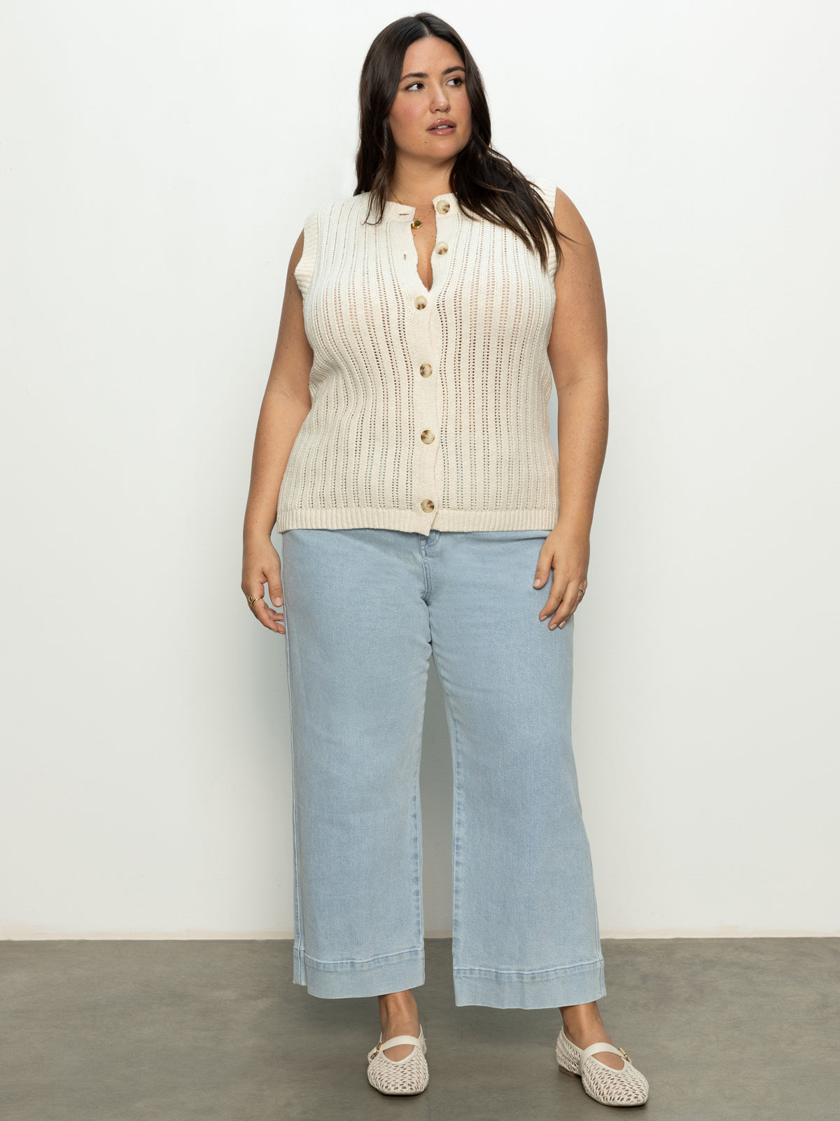 A person with long dark hair stands on a concrete floor against a plain white background, wearing the Sanctuary Clothing's pointelle shell eco natural inclusive collection top paired with light blue wide-leg jeans and woven slip-on shoes.