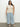 A person with long dark hair stands on a concrete floor against a plain white background, wearing the Sanctuary Clothing's pointelle shell eco natural inclusive collection top paired with light blue wide-leg jeans and woven slip-on shoes.