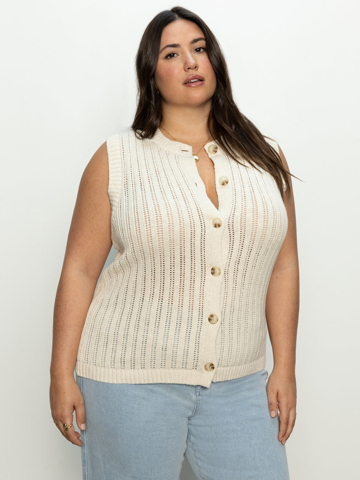 A woman with long dark hair is wearing Sanctuary Clothing's "pointelle shell eco natural inclusive collection" sleeveless, button-up beige knit top and light blue jeans, posing against a plain, light-colored background.