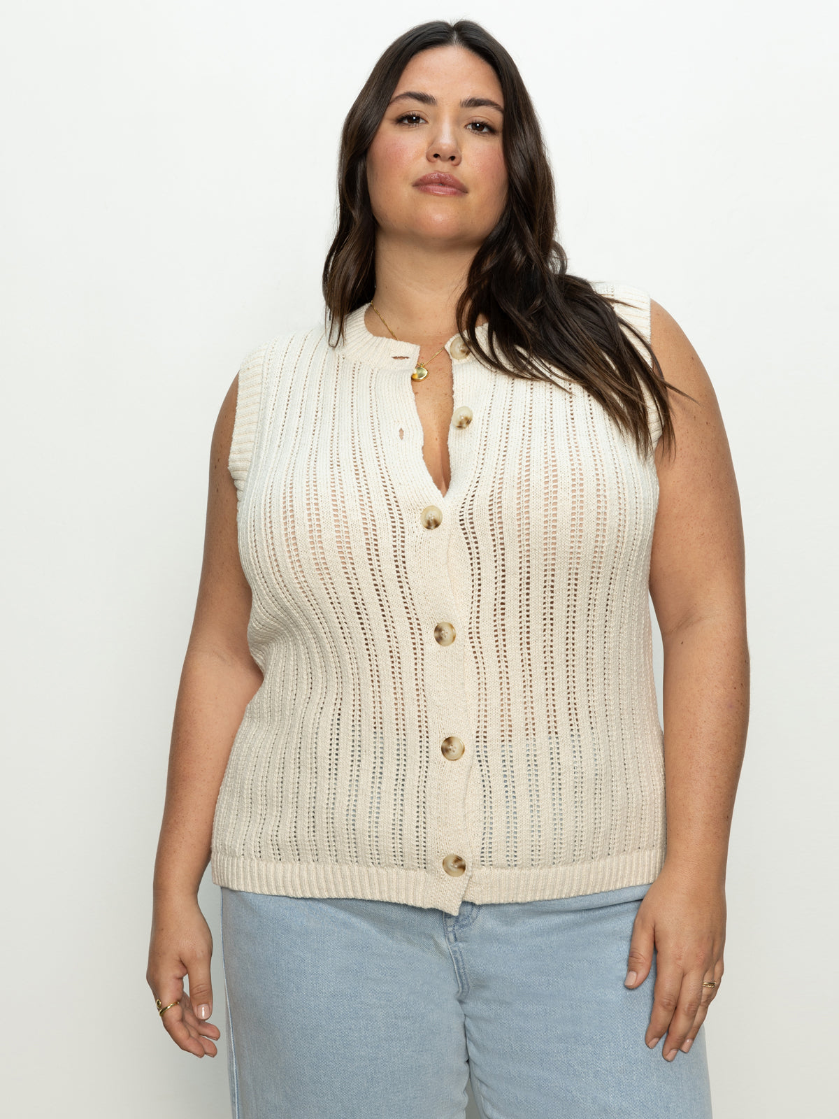 A person with long dark hair is wearing the Sanctuary Clothing 'pointelle shell eco natural inclusive collection' knit vest paired with light blue jeans. They stand against a plain light background, looking confidently at the camera.