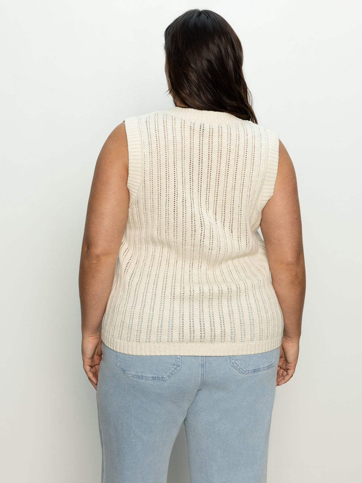 A person with long dark hair faces away from the camera, wearing Sanctuary Clothing's pointelle shell eco natural top from their inclusive collection and light blue jeans. The background is plain and light-colored.