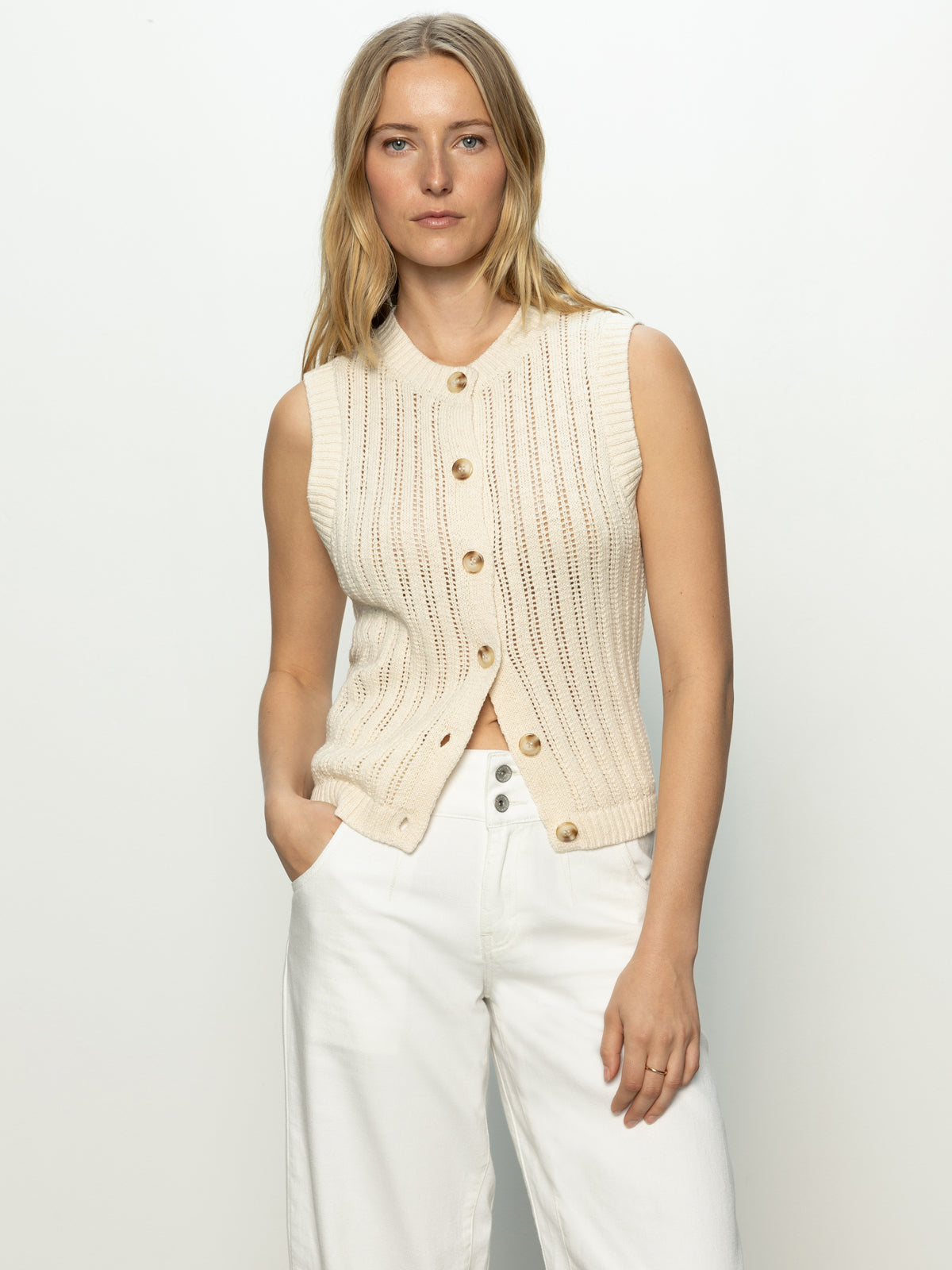 A person with long blonde hair wears the "Pointelle Shell Eco Natural" by Sanctuary Clothing, a sleeveless knit sweater with large buttons. Paired with white pants, they stand against a plain white background, one hand in their pocket.