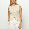 A person with long blonde hair wears the "Pointelle Shell Eco Natural" by Sanctuary Clothing, a sleeveless knit sweater with large buttons. Paired with white pants, they stand against a plain white background, one hand in their pocket.