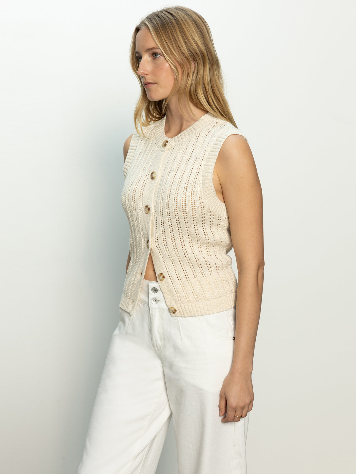 A person with long blonde hair is wearing the Pointelle Shell Eco Natural, a sleeveless knit top by Sanctuary Clothing, and white pants. They stand against a plain, light background, looking to the side.