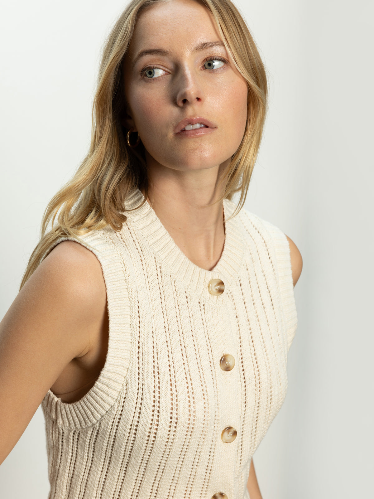 A woman with long blonde hair is wearing Sanctuary Clothing's "Pointelle Shell Eco Natural," a sleeveless, ribbed knit top with buttons. She gazes off to the side against a neutral background.