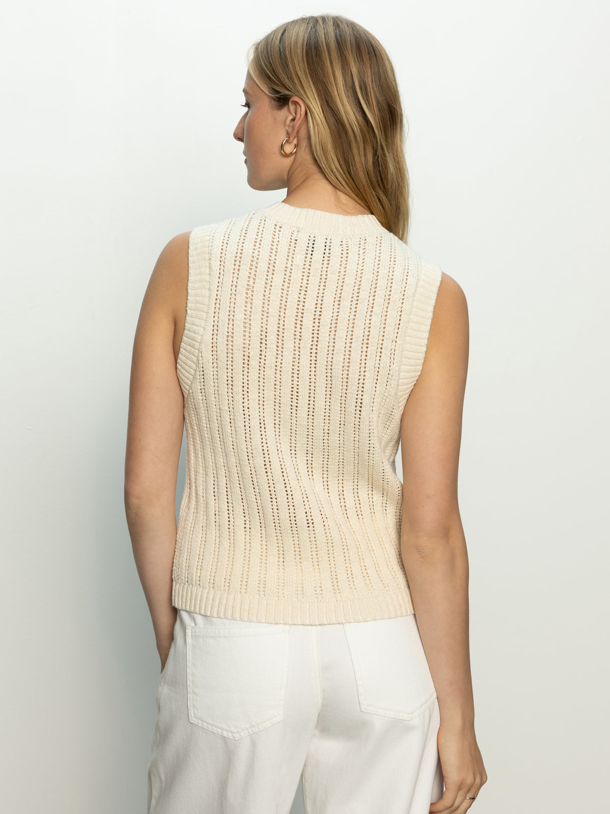 A woman with long blonde hair is shown from behind wearing Sanctuary Clothing's "Pointelle Shell Eco Natural," a sleeveless cream-colored knit sweater with vertical patterns. She has her hands in the pockets of her white pants against a simple, light backdrop.