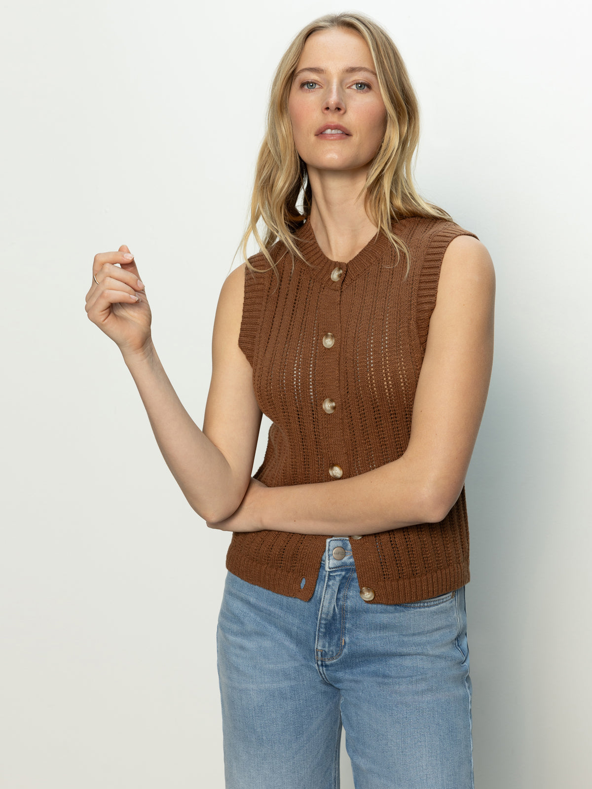 A woman with long blonde hair stands against a plain white background, wearing Sanctuary Clothing's pointelle shell sepia, a sleeveless brown knitted top with buttons, paired with light blue jeans.