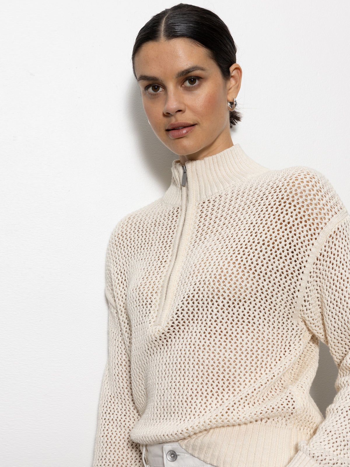 A dark-haired person wears the Sanctuary Clothing's "open knit 1/2 zip sweater eco natural," a light beige knitted sweater with a zipper, against a white background. The sweater features a textured, open-knit design as they face the camera.