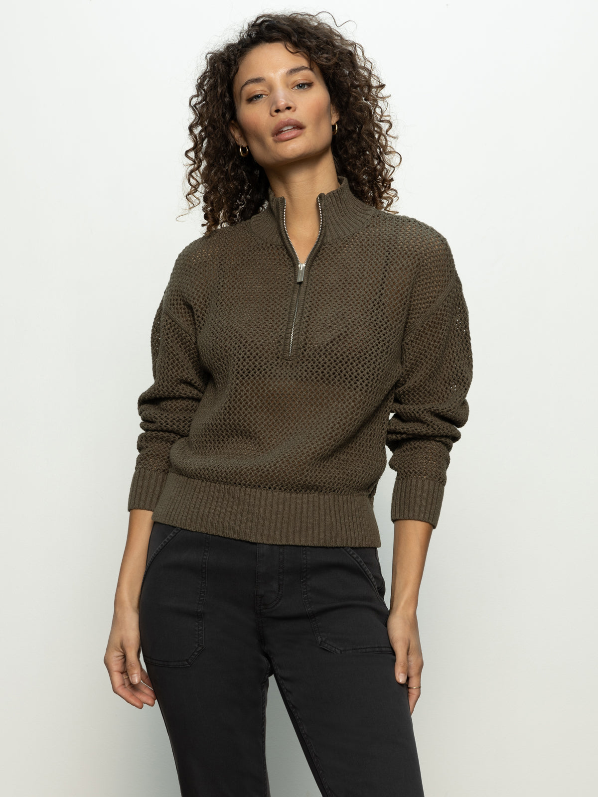 A person with curly hair is wearing Sanctuary Clothing's open knit 1/2 zip sweater in kalamata and dark jeans, standing against a plain light background.