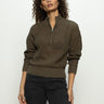 A person with curly hair is wearing Sanctuary Clothing's open knit 1/2 zip sweater in kalamata and dark jeans, standing against a plain light background.