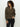 A person with curly hair is wearing a brown "open knit 1/2 zip sweater kalamata" by Sanctuary Clothing and black pants. They stand against a plain background, looking down with one hand touching their sweater.