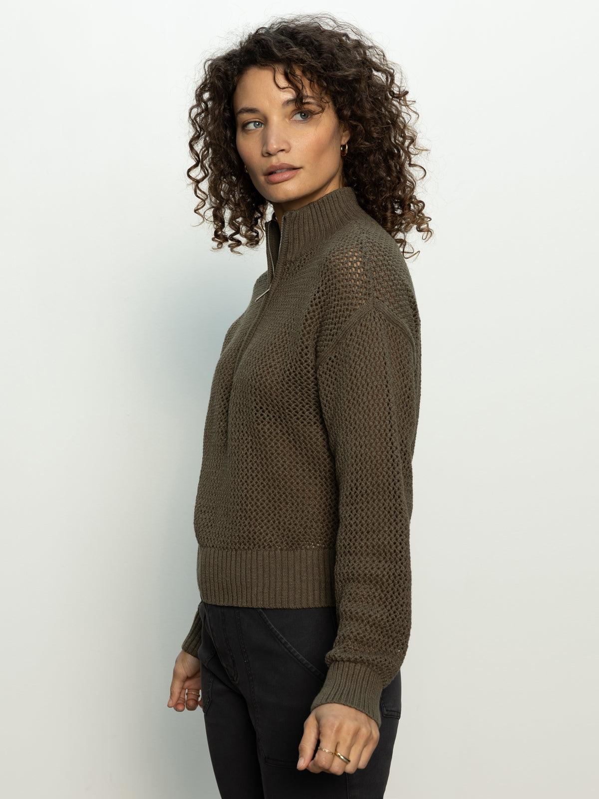 A woman with curly hair poses against a light background, looking to the side. She's wearing an open knit 1/2 zip sweater in kalamata from Sanctuary Clothing paired with dark pants.