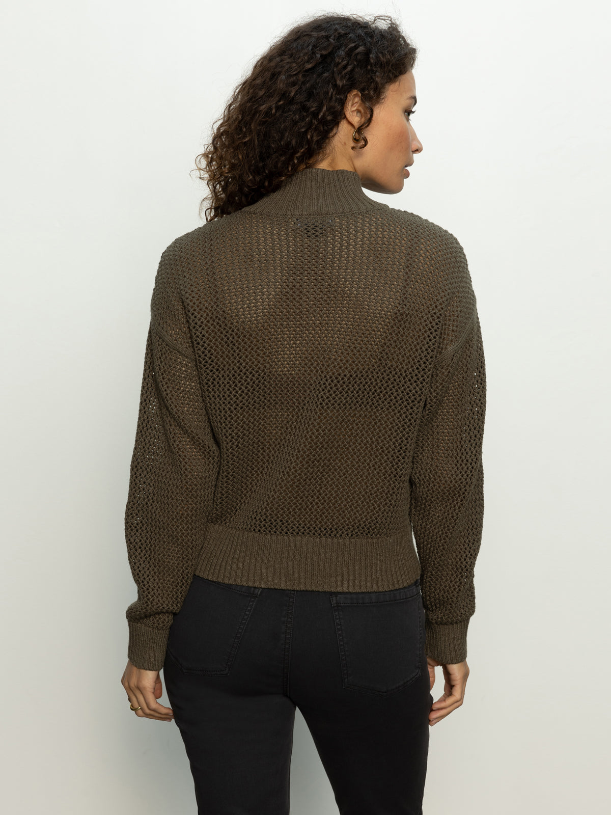 A person with curly hair is wearing a Kalamata open knit 1/2 zip sweater by Sanctuary Clothing and black pants. They are facing away, slightly turned to the side, against a plain light background.