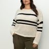 A person in a Sanctuary Clothing sporty stripe open knit sweater, featuring eco natural with black stripes from the inclusive collection, stands against a plain background. They have long brown hair and are wearing olive green pants, one hand in their pocket.