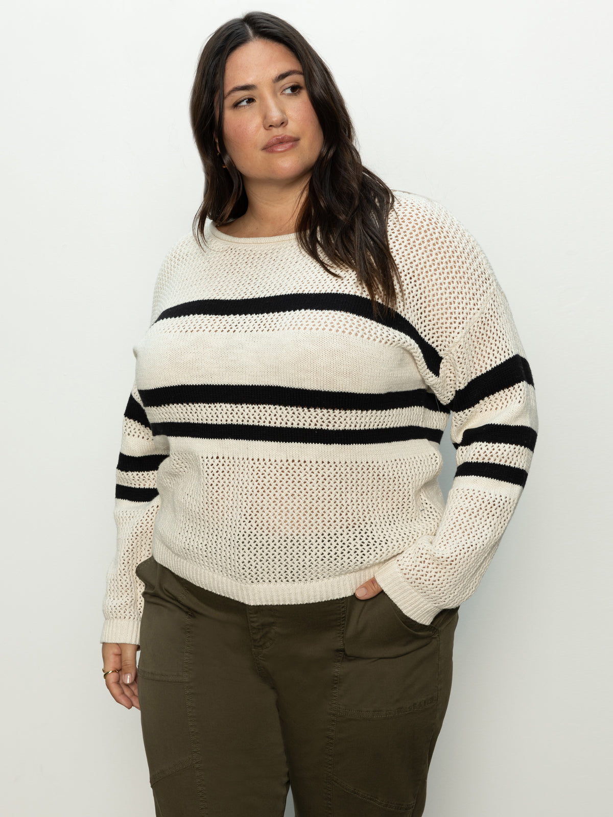 A person with long dark hair is wearing Sanctuary Clothing's sporty stripe open knit sweater in eco natural black stripe from the inclusive collection, paired with olive green pants. Against a plain light background, they gaze slightly to the side with a neutral expression.