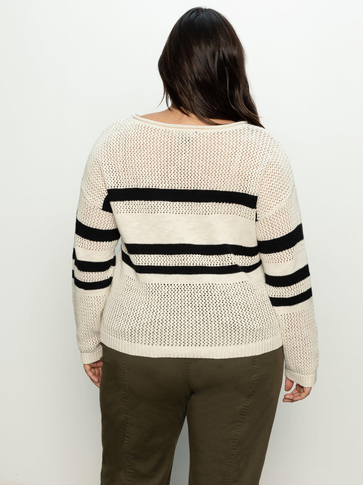 A person wearing Sanctuary Clothing's Sporty Stripe Open Knit Sweater in Eco Natural with Black Stripes from the Inclusive Collection is seen from the rear, paired with olive green pants. The background is plain white.