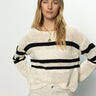 A person with long blonde hair wears the "sporty stripe open knit sweater eco natural black stripe" by Sanctuary Clothing, paired with light-colored pants, confidently posing against a neutral background.