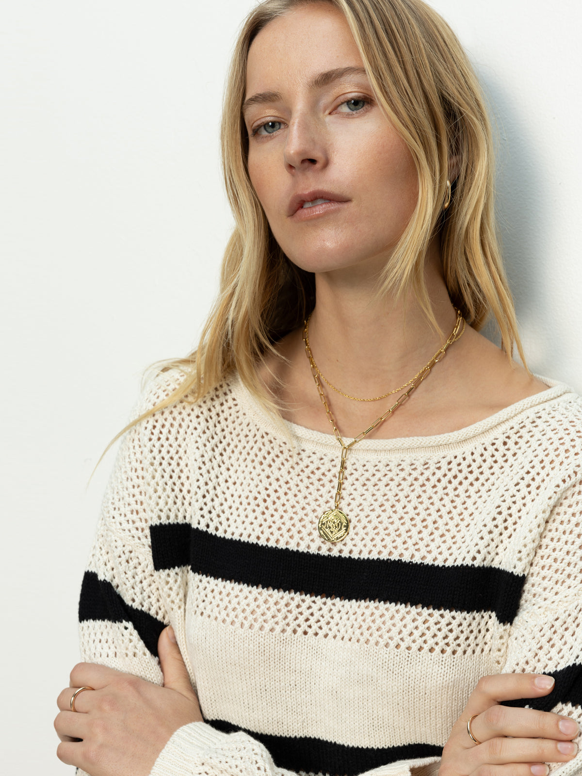 A woman with long blonde hair stands against a plain background in a Sanctuary Clothing sporty stripe open knit sweater in eco natural black stripe. She's wearing a gold necklace with a round pendant and has a calm expression, arms crossed casually.