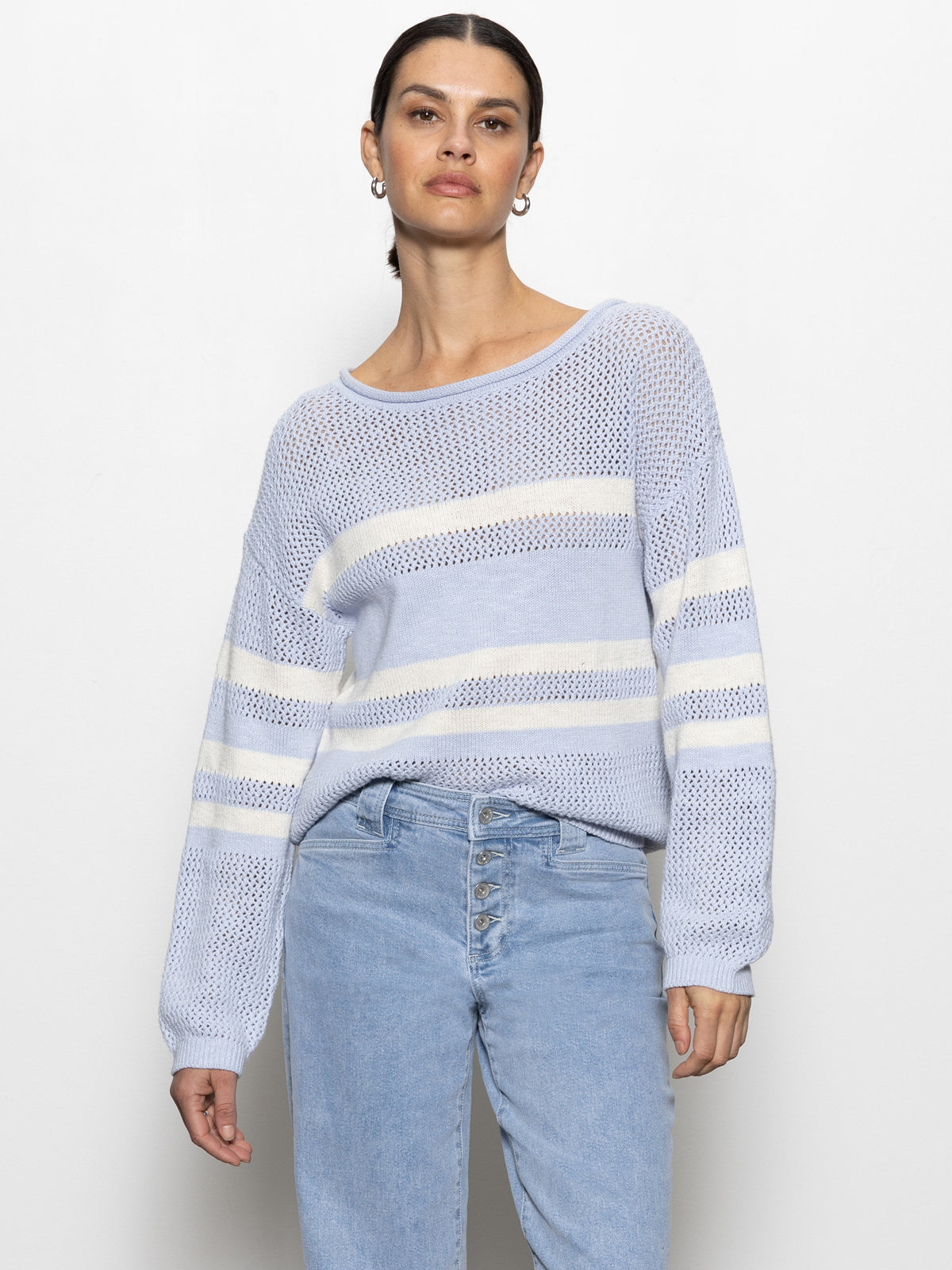 A person wearing the Sanctuary Clothing sporty stripe open knit sweater in sky blue with chalk stripes and light blue jeans stands against a plain white background.