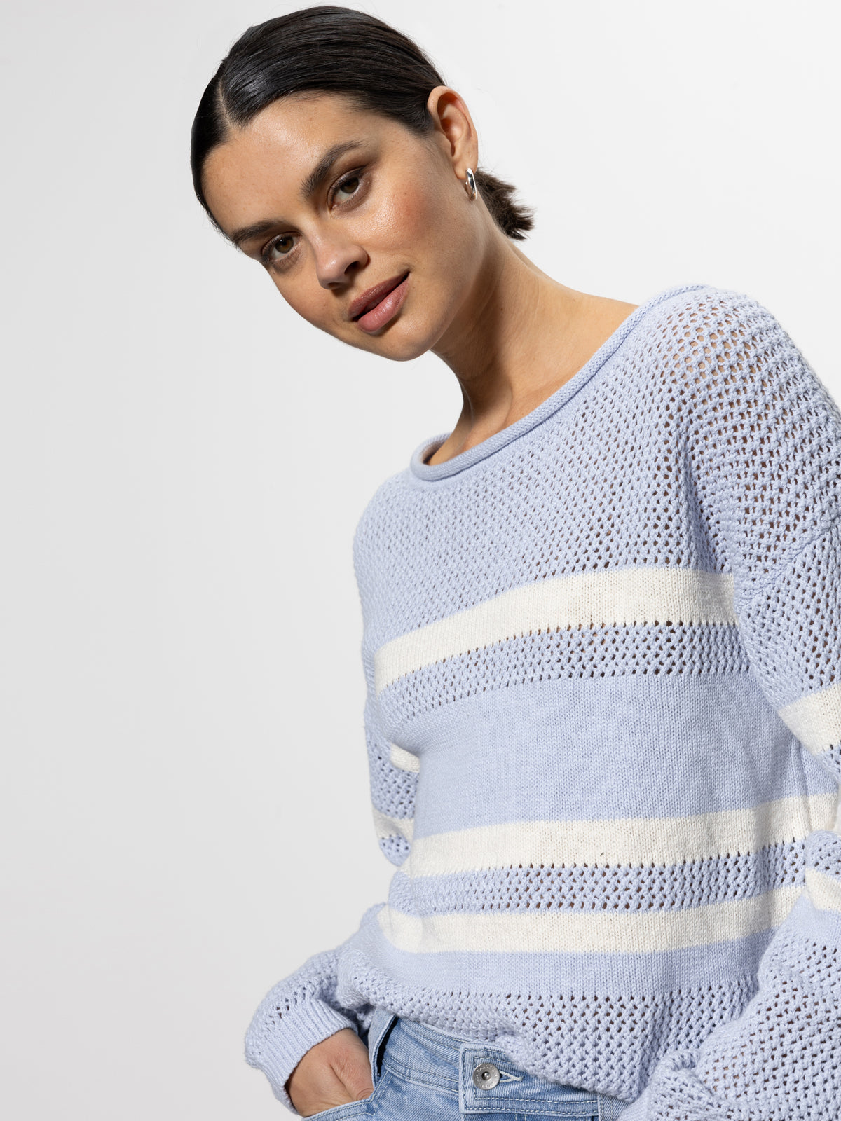 A person with dark hair pulled back is smiling slightly at the camera, wearing Sanctuary Clothing's sporty stripe open knit sweater in sky blue with chalk stripes. The background is plain and light-colored.