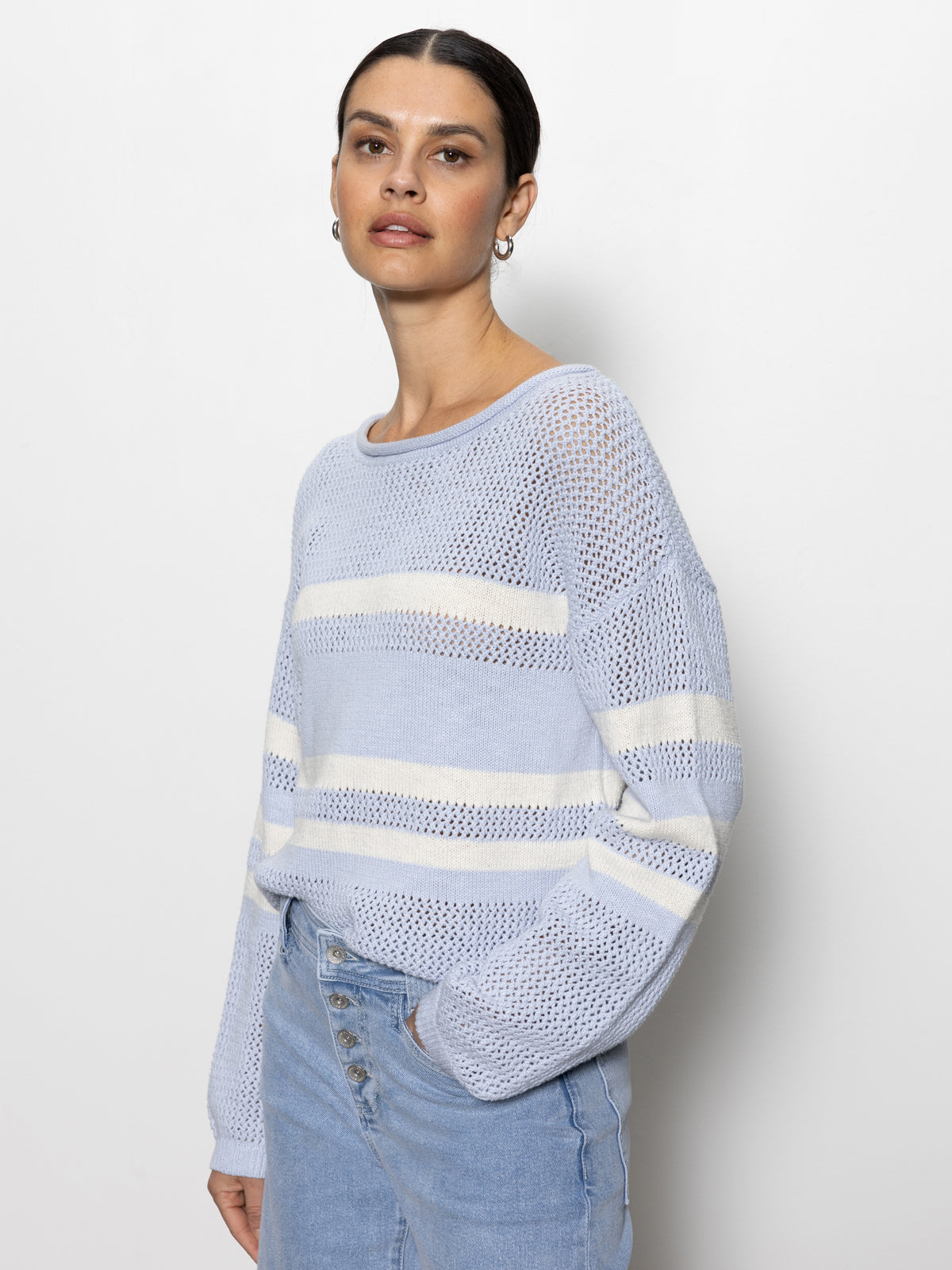 A person wearing a Sanctuary Clothing "sporty stripe open knit sweater" in sky blue with chalk stripes stands against a plain background. Their hands are in the pockets of light denim jeans, and they gaze at the camera with a neutral expression.