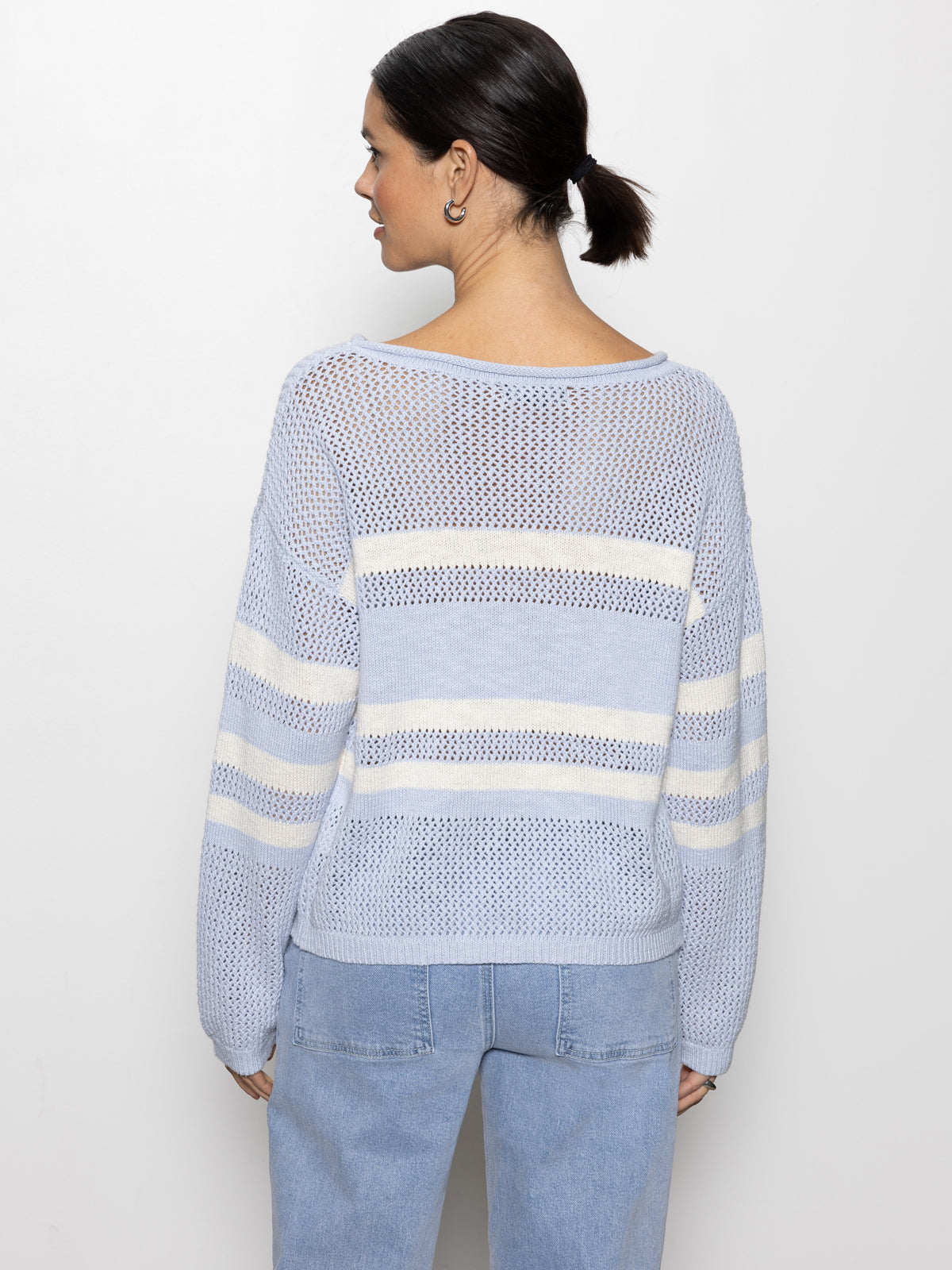 A woman with dark hair tied back wears a sporty stripe open knit sweater in sky blue with chalk stripe by Sanctuary Clothing and light blue jeans, standing against a plain white background and facing away.