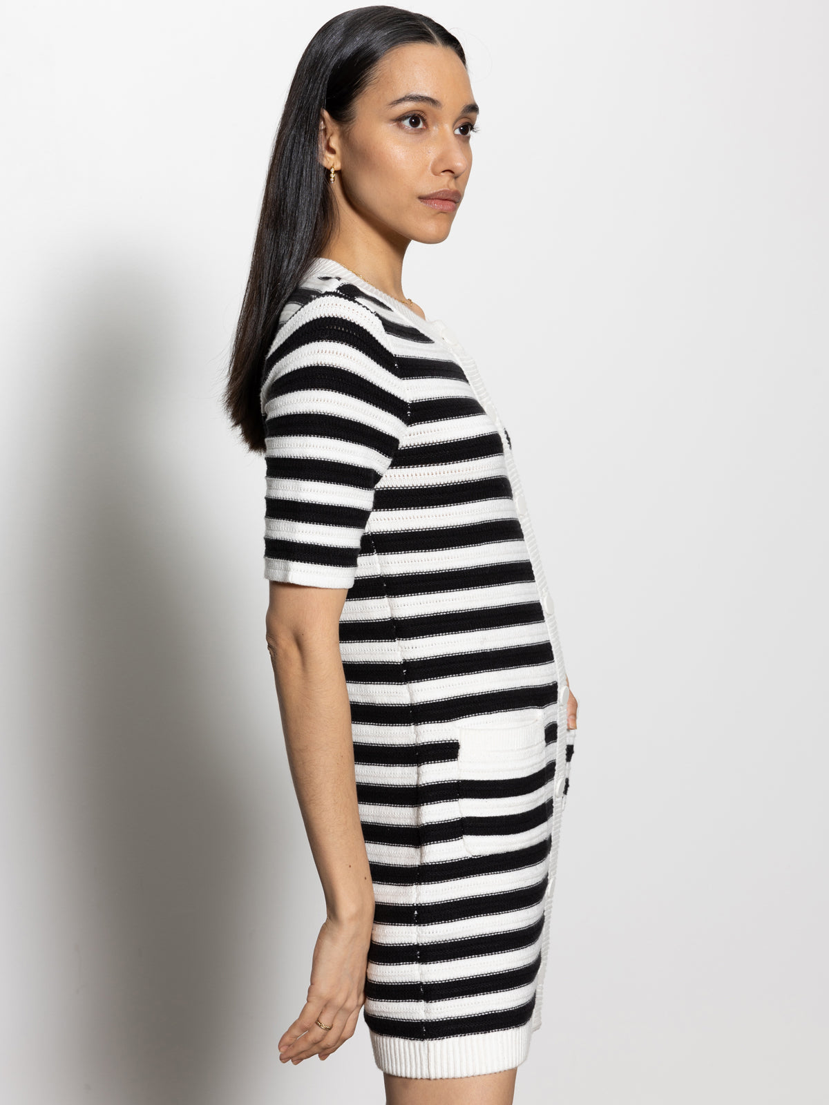 A person with long dark hair stands in profile against a white background, wearing the Sanctuary Clothing button-front knitted dress in chalk and black stripes, featuring short sleeves and pockets.