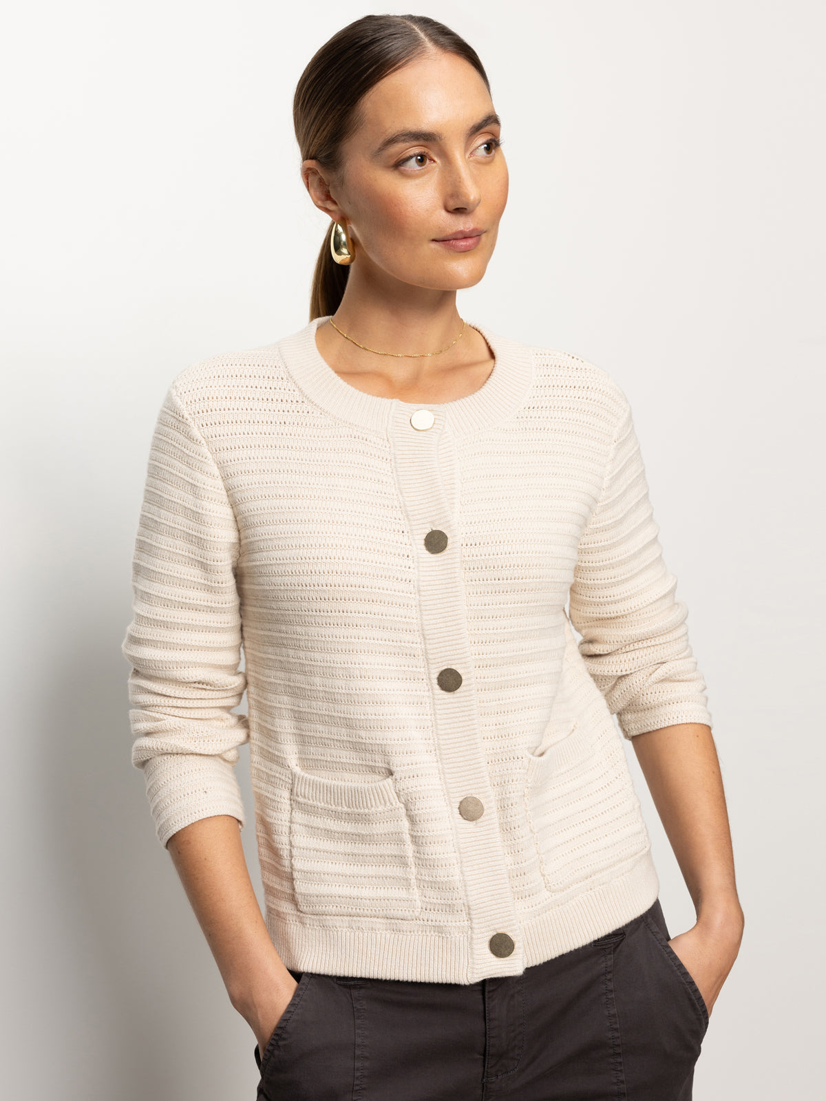 A person with long hair in a ponytail is wearing Sanctuary Clothing's pointelle knitted jacket in oat, paired with dark pants. The cream-colored jacket features two pockets and large buttons. They are looking to the side against a plain background.