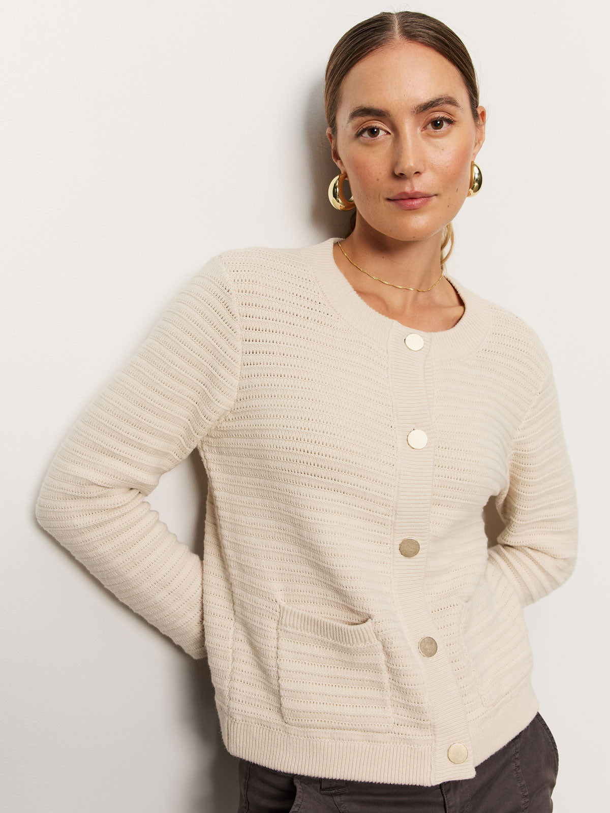 A person with long hair, wearing large hoop earrings and a cream-colored pointelle knitted jacket by Sanctuary Clothing, poses against a light-colored wall. The jacket features large buttons, two front pockets, and textured horizontal stripes.