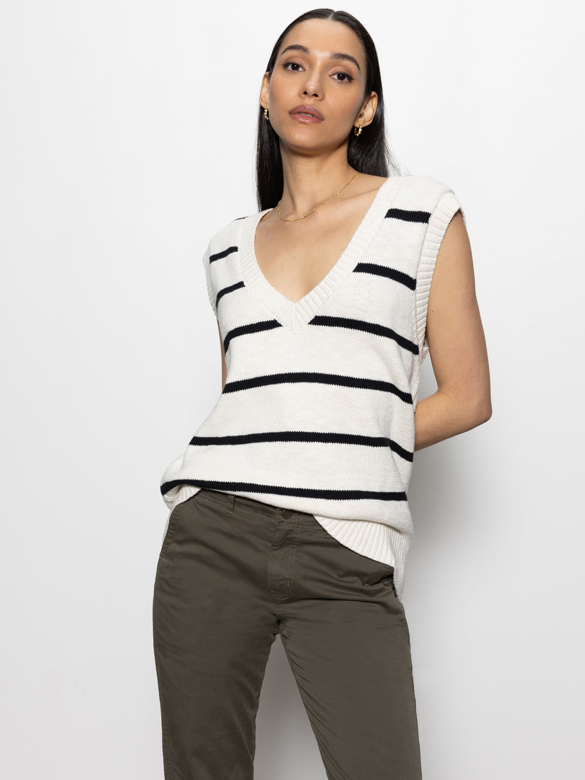 A person with long black hair is wearing a Sanctuary Clothing bf sweater vest in chalk with black, paired with olive green pants. They are posing against a plain white background.