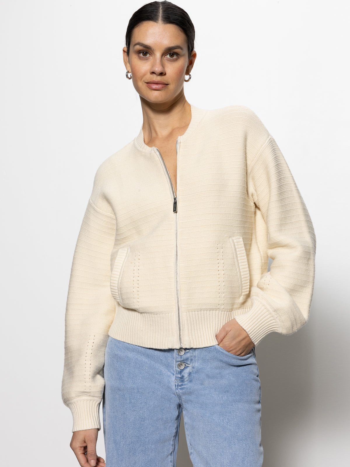 A person is wearing the "knitted bomber muslin" by Sanctuary Clothing, a cream-colored zip-up knit sweater with large pockets, paired with light blue jeans. Their hand is in one pocket as they stand against a plain white background, looking at the camera.