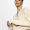 A woman wearing a Sanctuary Clothing "knitted bomber muslin" with a full zipper poses against a white background. She has dark hair pulled back, hoop earrings, and gently clasped hands by her face.