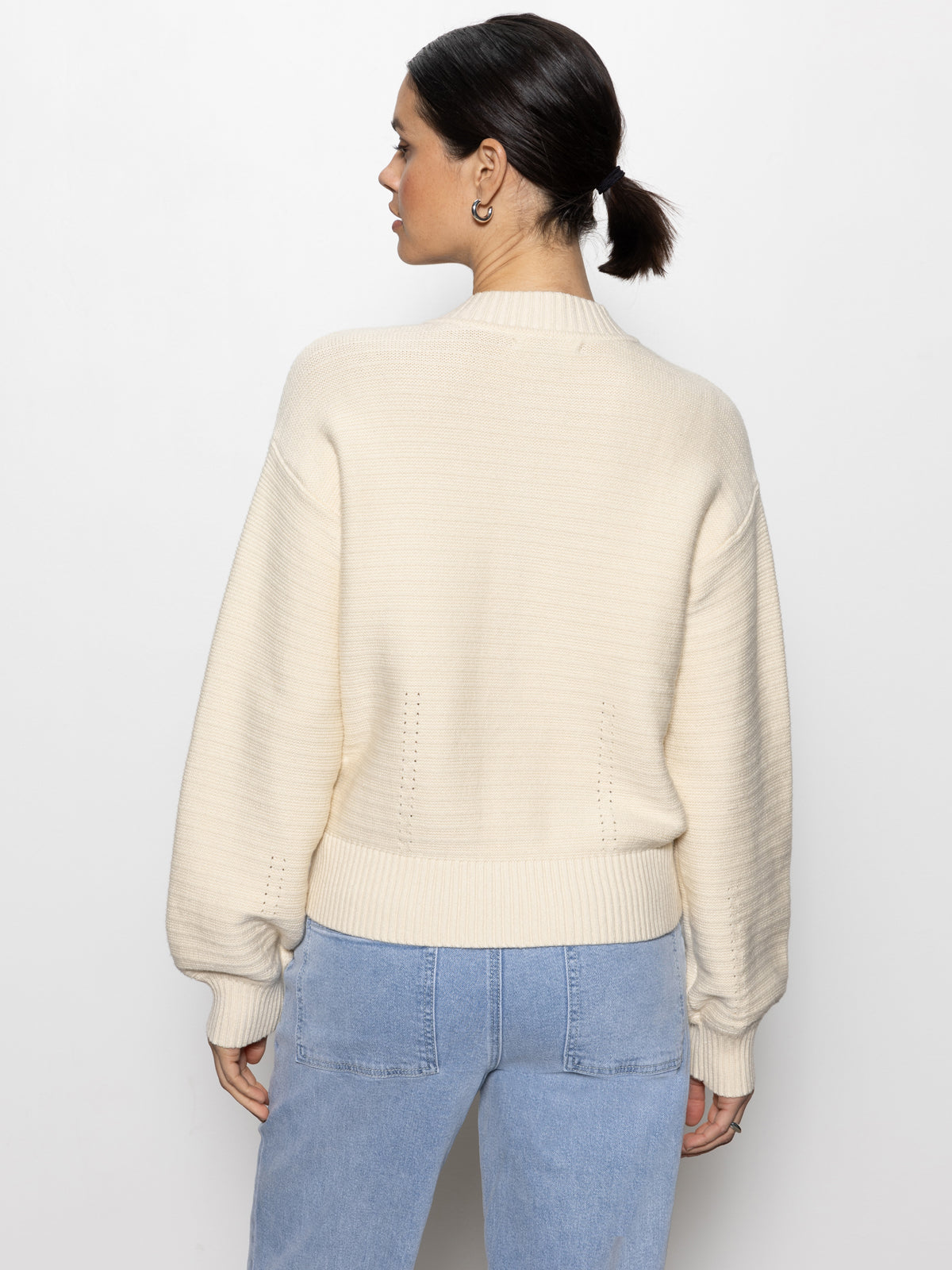 A person with dark hair in a ponytail stands with their back to the camera, wearing Sanctuary Clothing's cream-colored knitted bomber muslin and light blue jeans. The backdrop is plain white.