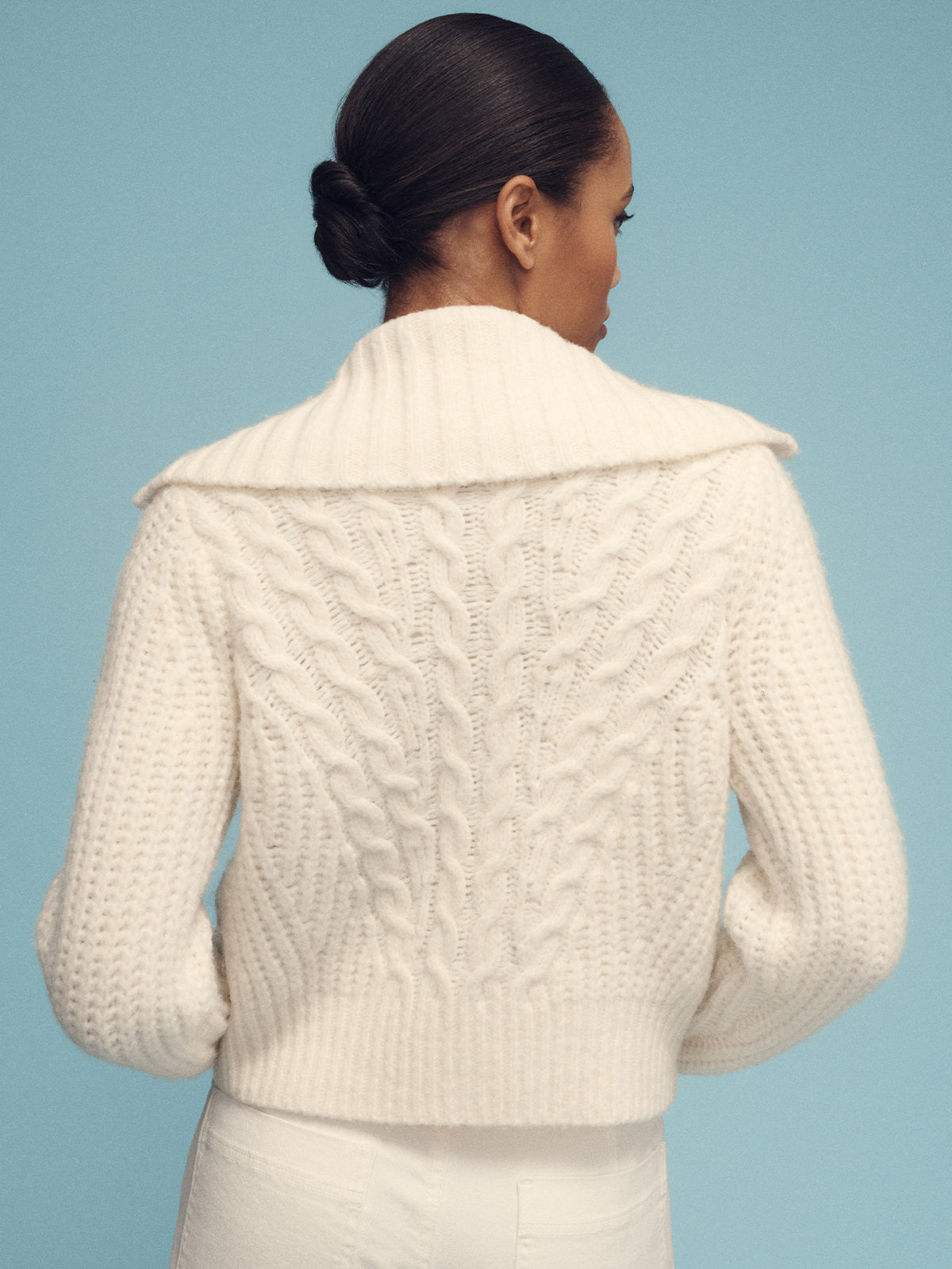 A person with dark hair in a bun is wearing the Sanctuary Clothing cable zip sweater jacket in chalk, characterized by its chunky cable-knit design and wide collar. They are facing away against a light blue background.